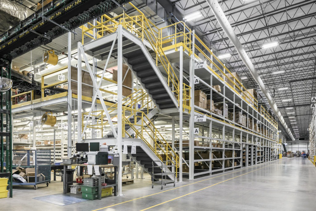Industrial warehouse interior with multi-level steel shelving, stairs, and conveyor systems.